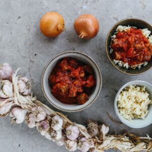 Top view of a rustic meal with garlic, onions, and bowls of rice and stew on a stone surface.